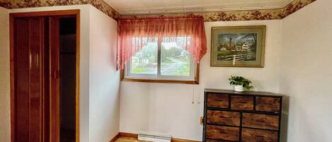 Una habitación con piso de madera, una ventana con cortina rosa, un cuadro en la pared y un mueble de madera.