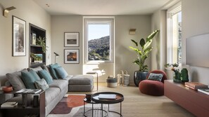 Een moderne woonkamer met een grijze bank, een rode poef, een ronde salontafel en een groot raam met uitzicht op heuvels.