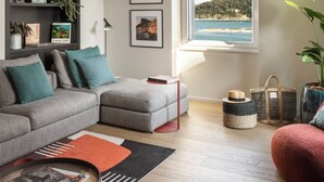 A modern living room with a grey sofa, a red ottoman, a coffee table, a bookshelf, and a large window with a view of the sea. - San Pietro Terrace by the Sea (Portovenere)