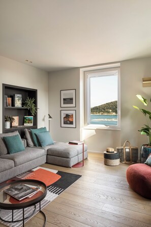 A modern living room with a grey sofa, a red ottoman, a coffee table, a bookshelf, and a large window with a view of the sea. - SAN PIETRO TERRACE BY THE SEA Seafront Luxury Terrace Apartment - Portovenere (Portovenere)