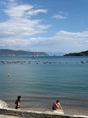 A coastal scene with a pebbled beach, calm sea, boats, and distant hills. - SAN PIETRO TERRACE BY THE SEA Seafront Luxury Terrace Apartment - Portovenere (Portovenere)