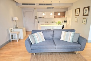 A modern living room with a grey sofa, a small white table, and a lamp. A kitchen is visible in the background with white cabinets and pendant lights. - 1 Bed Serviced Panoramic SkyVilla (Krung Thep Maha Nakhon)