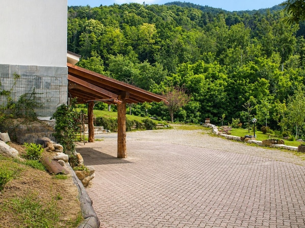 Un sendero pavimentado que conduce a un edificio con estructura de pérgola, rodeado por una ladera boscosa.