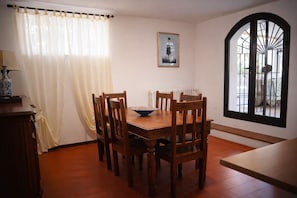 A dining area with a wooden table, chairs, a bowl of food, a window with bars, and a radiator.