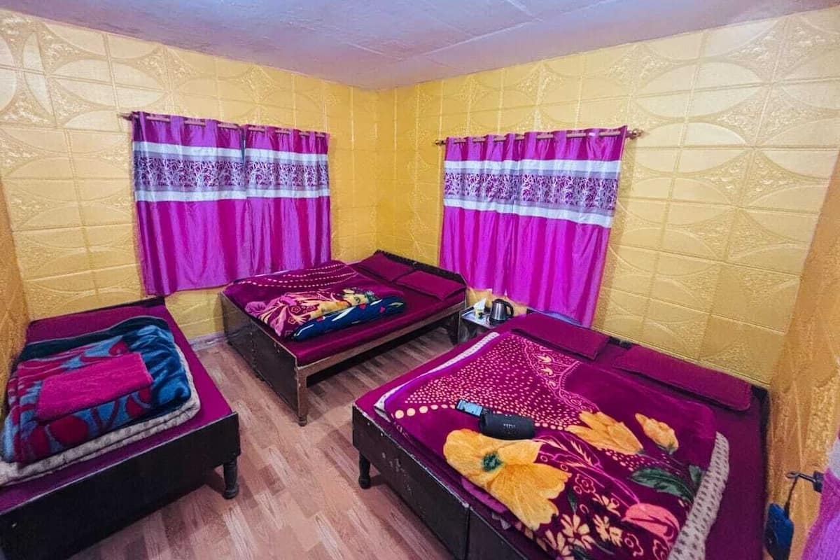 Quarto com três camas, cada uma com colcha de padrão diferente, e duas janelas com cortinas roxas.