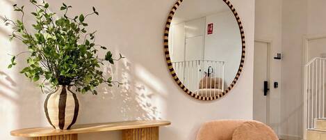 A room with a wooden console table, a round mirror with a striped frame, a plush armchair, and a potted plant.