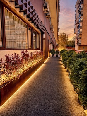 Passadís amb un edifici modern a un costat, un camí i una filera de plantes il·luminades per llums.