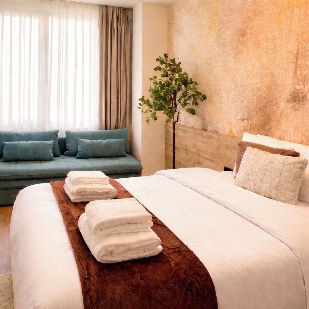 Una habitación de hotel con cama, sofá, planta y ventana con cortinas.