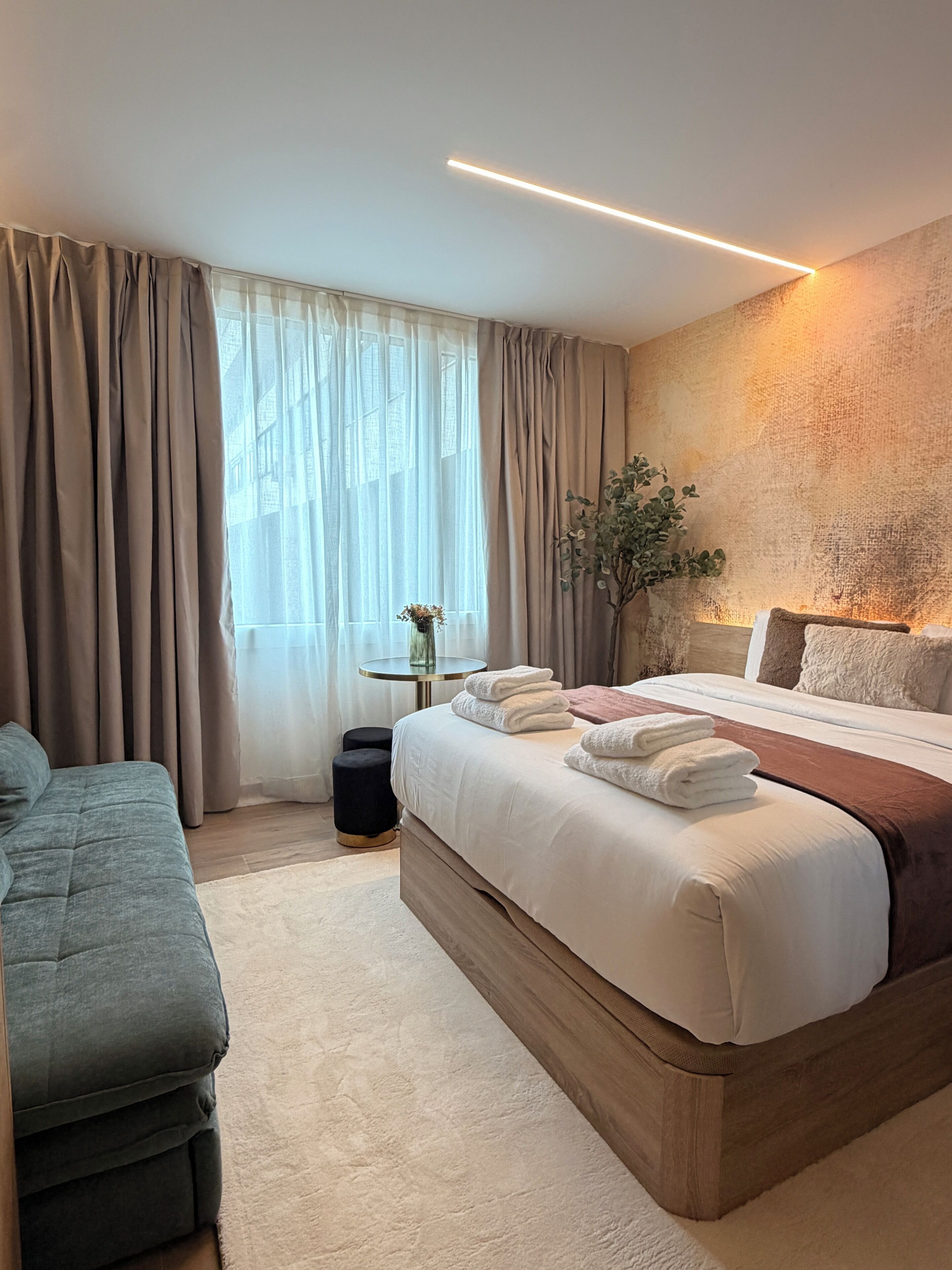 A modern bedroom with a large bed, a bedside table with a vase, and a view of the outside through sheer curtains.