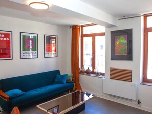 Un salón con un sofá azul, una mesa de centro de cristal y un radiador. Hay obras de arte enmarcadas en la pared y una ventana con cortinas.