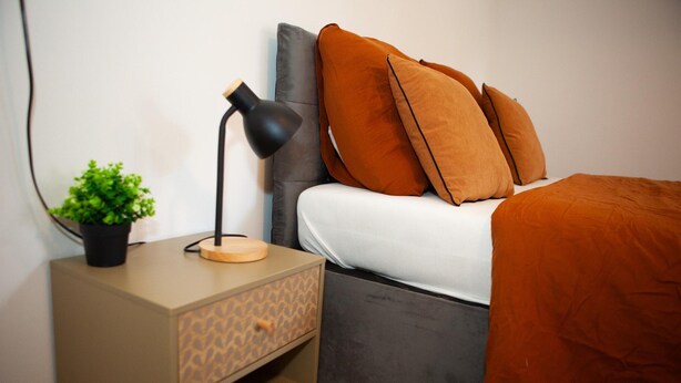 Ein Bett mit grauem Kopfteil, orangefarbener Bettwäsche, einer Nachttischlampe, einem kleinen Topfpflanzen und einer Nachttischschublade mit Muster.