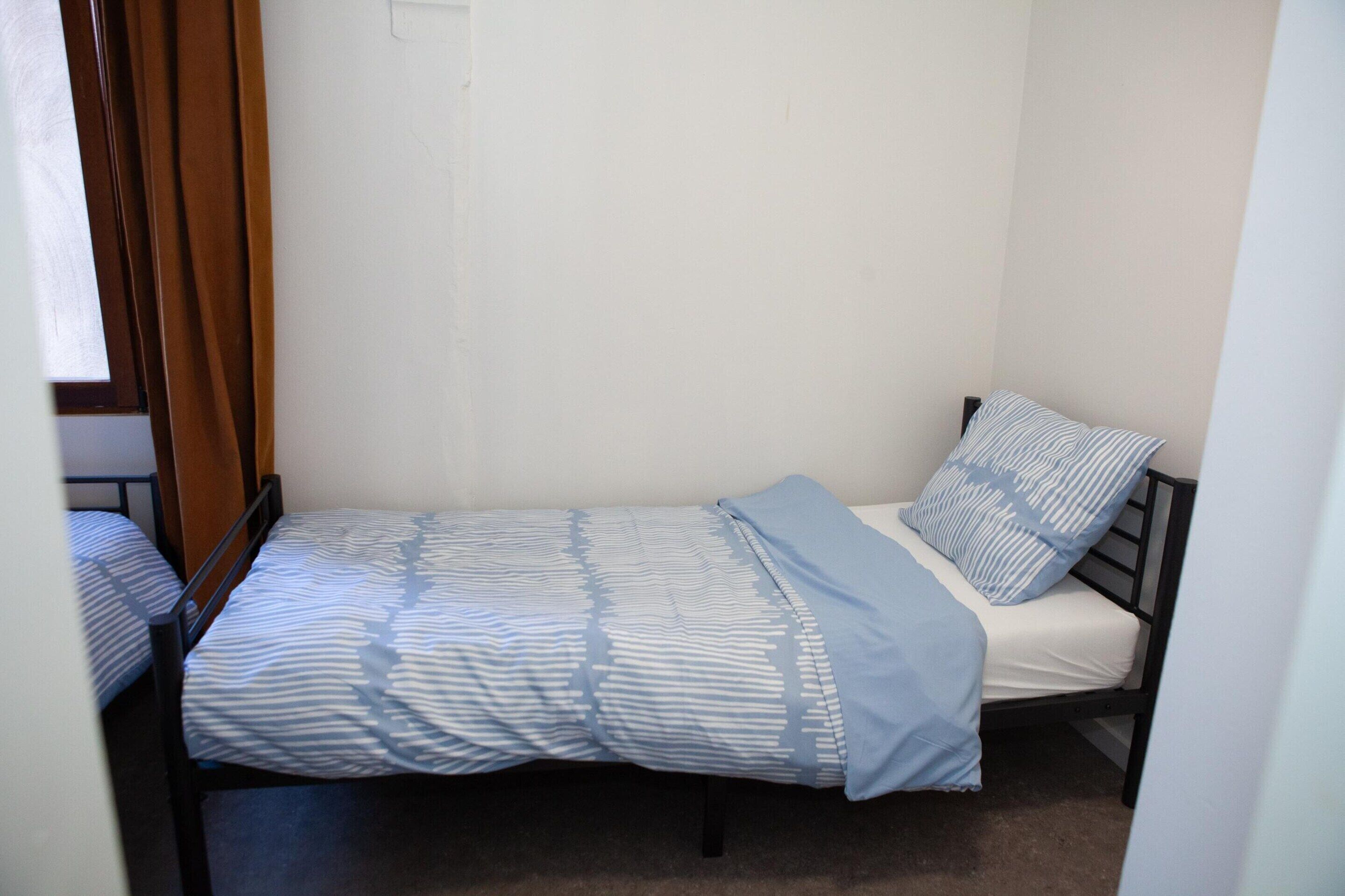 A single bed with a striped bedspread and pillows, a wooden door frame, and a plain wall.