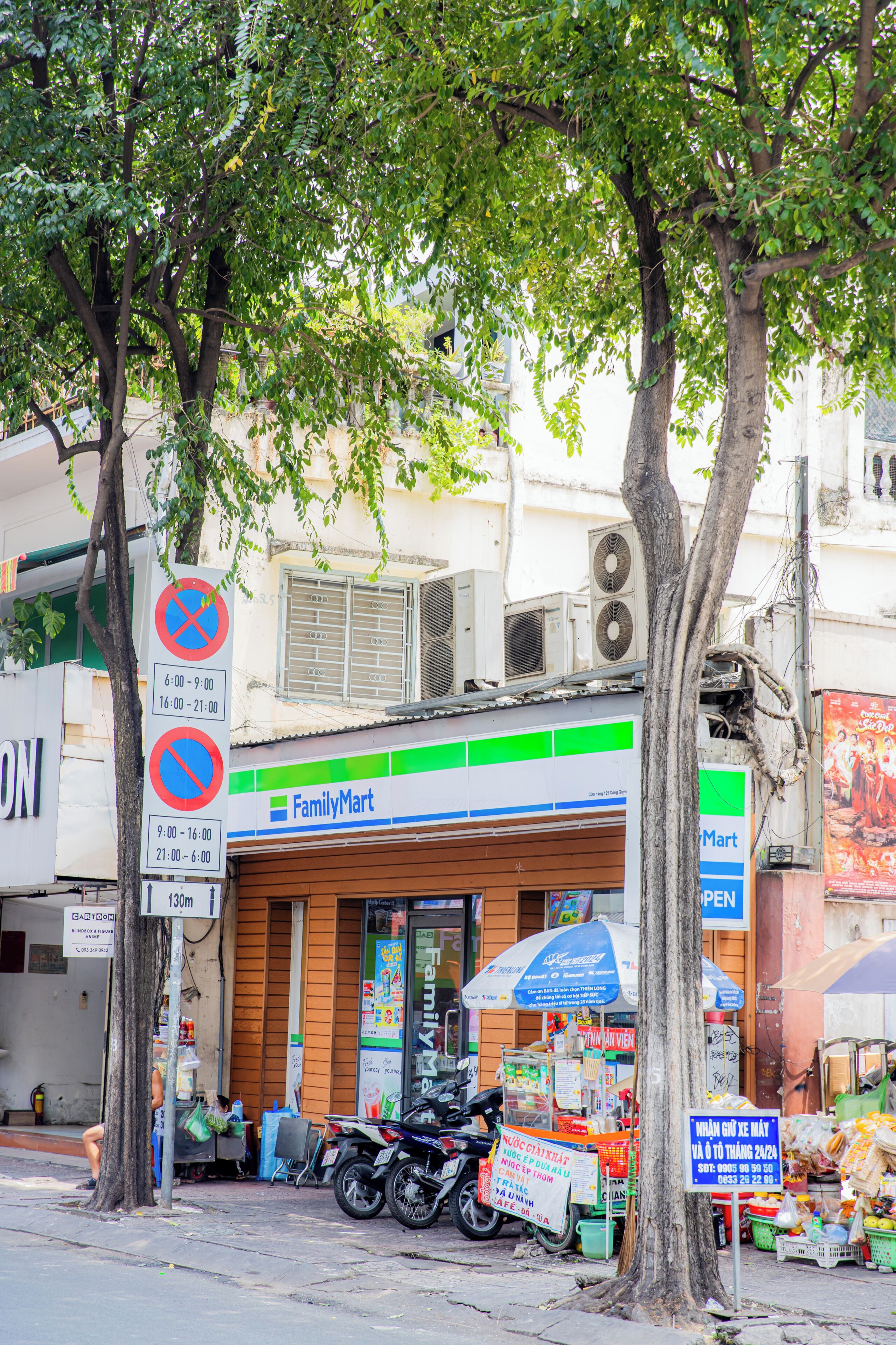 Una tienda FamilyMart con una motocicleta estacionada frente, un letrero que indica prohibido estacionar y un árbol al frente.