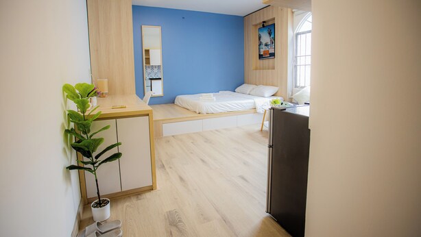 Quarto compacto com cama, escrivaninha, geladeira, planta em vaso e espelho.