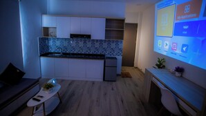 Cozinha moderna com armários brancos, painel de backsplash com padrão azul e forno embutido. Há uma pequena mesa com uma planta e um controle remoto, e uma tela de projeção exibindo ícones de aplicativos.