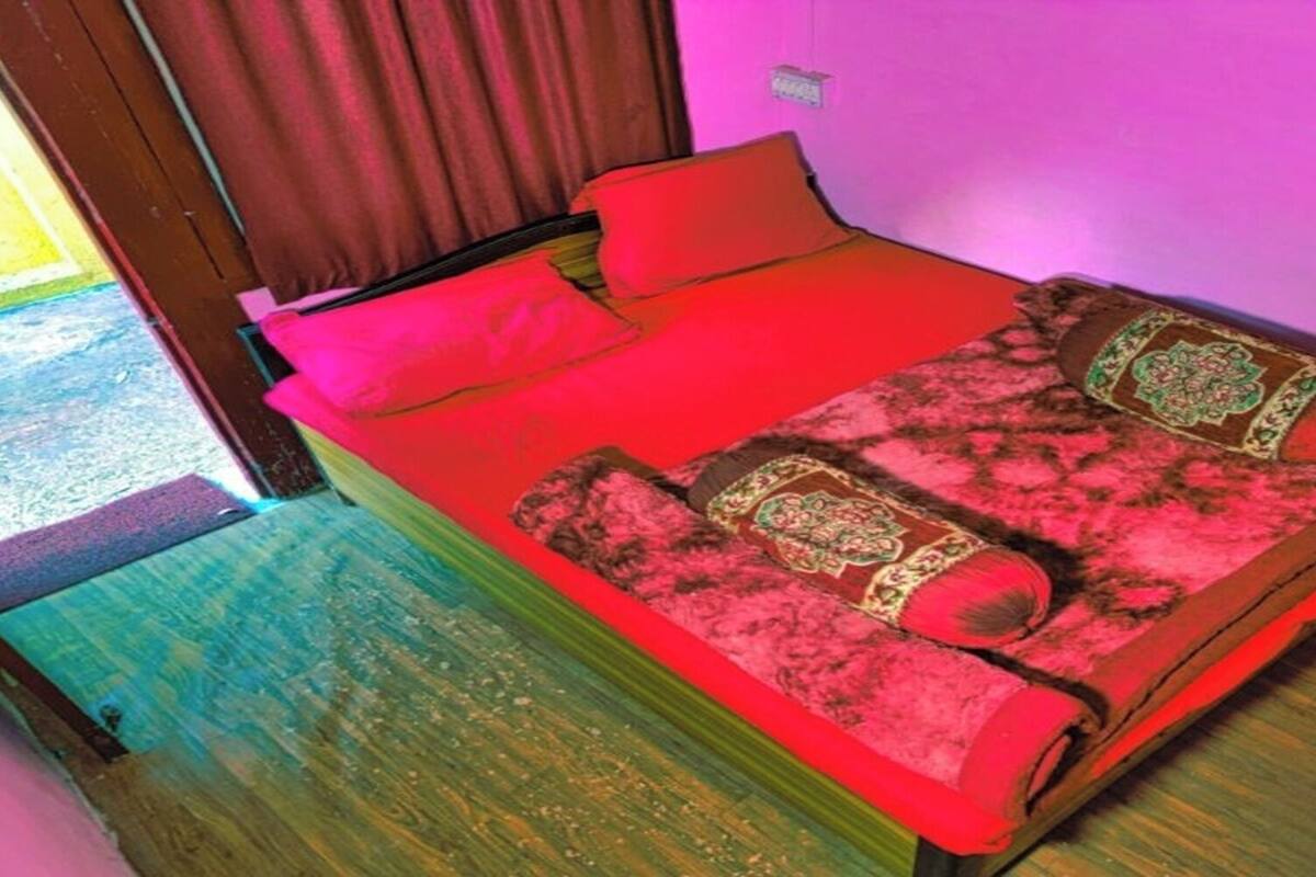 A single bed with a red blanket and pillows, a wooden floor, and a pink wall.