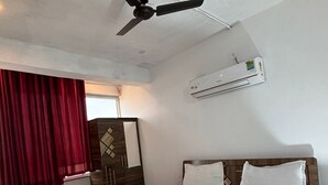 Et soveværelse med seng, ventilator, klimaanlæg, rødt gardin, et lille bord og et garderobeskab.