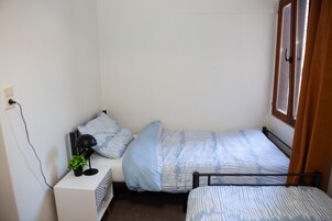 Habitación pequeña con cama individual, ventana, mesita de noche con lámpara y toma de corriente.