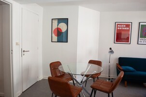 A room with a glass table surrounded by four chairs, a sofa, and two framed artworks on the wall.