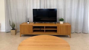 En stue med et tv-møbel i træ, et fladskærms tv, et rundt sofabord og to krukker med planter.