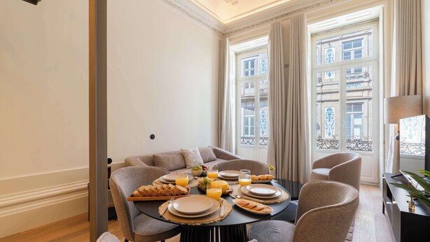 Ein Essbereich mit Tisch, gedeckt für das Frühstück, einschließlich Teller, Gläser und Brot. Stühle stehen um den Tisch, im Hintergrund ist eine Couch zu sehen.