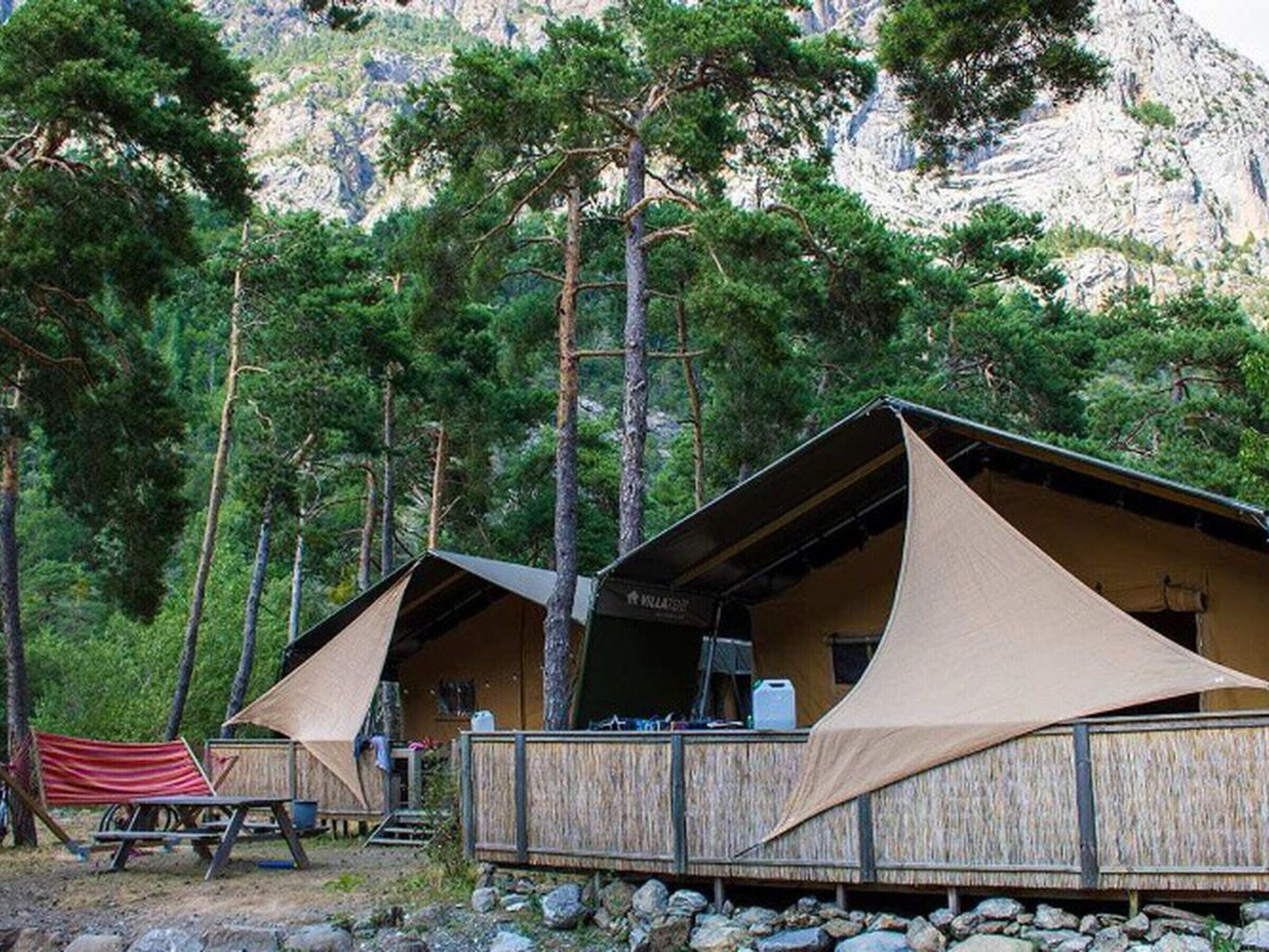 Deux cabanes en bois, dotées d’auvents en toile, entourées d’une forêt dense et d’un terrain rocheux.