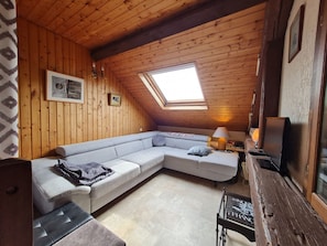 Une pièce mansardée chaleureuse avec un plafond en pente en bois, une grande fenêtre, un canapé d’angle, une petite table avec une lampe et un téléviseur.