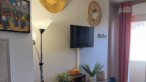 Et værelse med et ophængt fjernsyn, en hat på væggen, et indrammet billede, en lampe, en hylde med planter, og et skilt med teksten 'Bonjour'.
