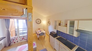 A compact kitchen with a sink, stove, and cabinets. There is a dining table with chairs and a clock on the wall.