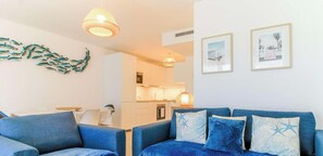 Ein Wohnzimmer mit einem blauen Sofa, einer Küche im Hintergrund und einer Wand mit gerahmten Kunstwerken.