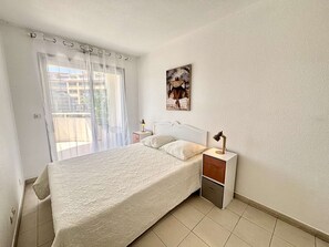A bedroom with a bed, a nightstand, a lamp, a window with sheer curtains, and a wall-mounted picture. - Bright apartment, park view - 4 guests (Juan les Pins)