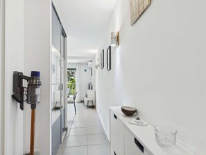 A hallway with a modern design, featuring a glass door, a wall-mounted vacuum cleaner, a white cabinet with a bowl and glass, and a framed picture on the wall. - Apartment with AC and WiFi - 5 guests - In the village center (Le Grau d'Agde)
