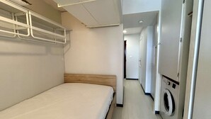 Ein schmaler Hotelkorridor mit einem Bett auf der linken Seite, einer Waschmaschine auf der rechten Seite und einer Tür am Ende.