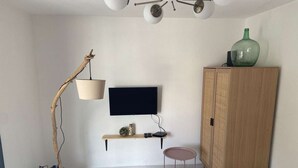 Ein Zimmer mit an der Wand befestigtem Fernseher, einem Holzschrank, einem runden Tisch und einer Stehlampe.