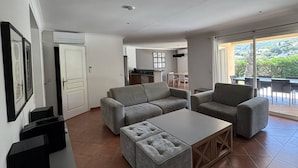 A modern living room with a sofa, ottoman, and coffee table, a kitchen in the background, and a balcony with a view. - Quiet villa with air conditioning and pool (Cavalaire-sur-Mer)