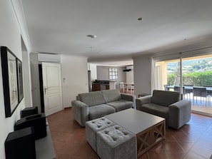 Ein modernes Wohnzimmer mit Sofa, Hocker und Couchtisch, im Hintergrund eine Küche und ein Balkon mit Aussicht.