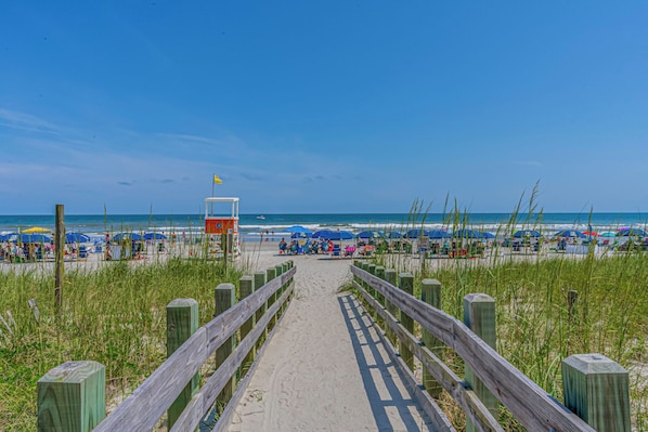Ein Strandweg führt zu einem Strandabschnitt mit Sonnenschirmen und Badegästen.