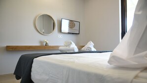 A neatly made bed with white linens, a round mirror, a framed picture, and a towel rack with towels.