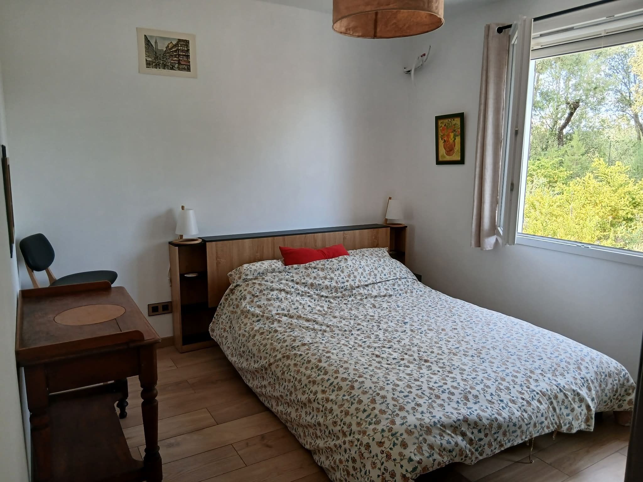Ett sovrum med en säng, ett skrivbord, en stol, en lampa och ett fönster med utsikt över träd.