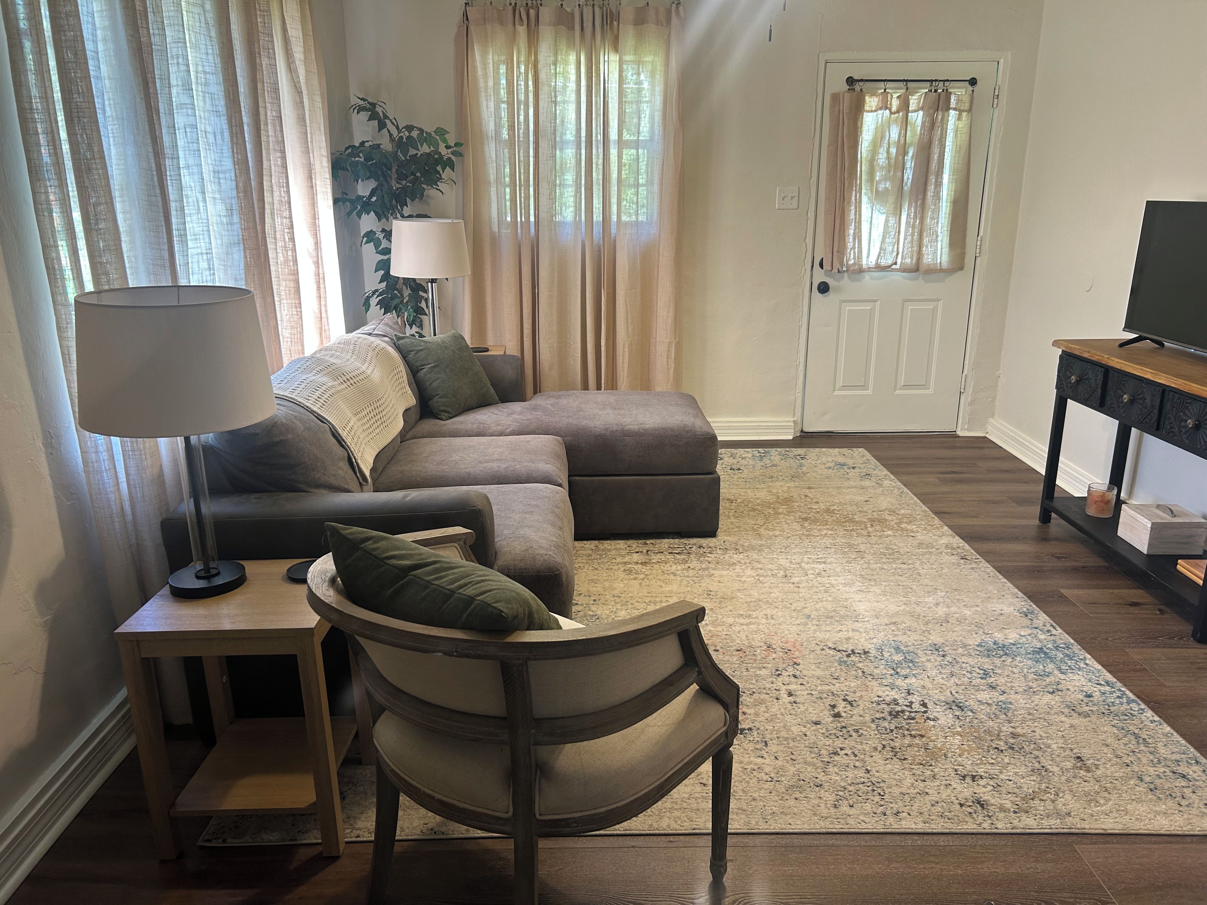 Un salon comprenant un canapé d’angle, une table d’appoint avec une lampe, une chaise en bois, un téléviseur posé sur un meuble et une porte avec des rideaux.