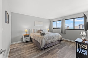 A bedroom with a bed, bedside tables, a window with curtains, and a view of buildings. - Seascape Cove at Holiday Towers 605 elevator/pool (Myrtle Beach)