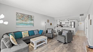 Ein modernes Wohnzimmer mit einem grauen Sofa, Sesseln und einem Couchtisch. Im Hintergrund ist eine Küche erkennbar.