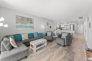 A modern living room with a grey sofa, armchairs, and a coffee table. A kitchen is visible in the background. - Seascape Cove at Holiday Towers 605 elevator/pool (Myrtle Beach)