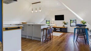 A modern kitchen with a central island, wooden stools, a TV mounted on the wall, and a view of the outdoors. - Ferny Retreat -  Romantic barn conversion, Hay-On-Wye (Bromyard)