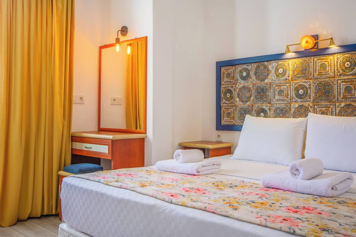 Habitación de hotel con cama, mesita de noche, espejo, escritorio de madera y cortina amarilla.