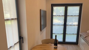 Ett rum med ett runt träbord, två vita stolar, en vinflaska, en tv på väggen och ett stort fönster med persienner.