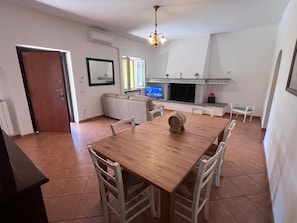 A dining area with a wooden table and white chairs, a television, a fireplace, and a door. - La casa di Marina Guest House (Monte Migliore-la Selvotta)