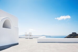 Ein weißes Gebäude mit einer gewölbten Fassade und einem flachen Dach, zwei weiße Stühle und Blick auf das Meer und ferne Berge.
