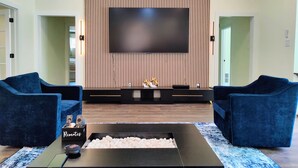Una sala de estar moderna con un televisor de pantalla plana grande montado en una pared de paneles de madera, dos sofás azules, una mesa de centro con un control remoto y un objeto decorativo pequeño en el mueble de la televisión.