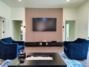 A modern living room with a large flat-screen TV mounted on a wooden panel wall, two blue sofas, a coffee table with a remote control, and a small decorative item on the TV stand. - 3-BDR Resort-style Home - Hot Tub, Mini Golf, Fire Pit, Grill, 1Gig WiFi, Office (Richardson)